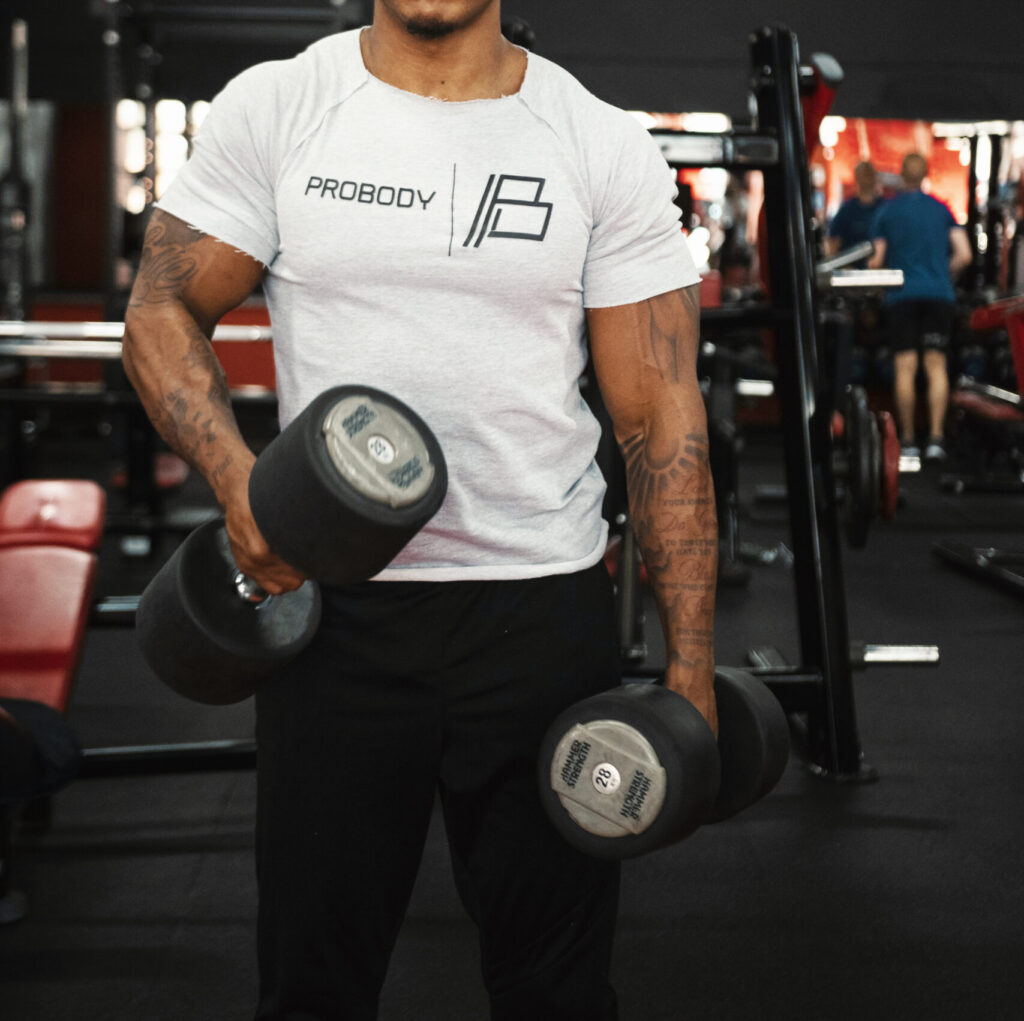 Man training with dumbbells in a gym, wearing a white ProBody T-shirt with the PROBODY B-mark chest print